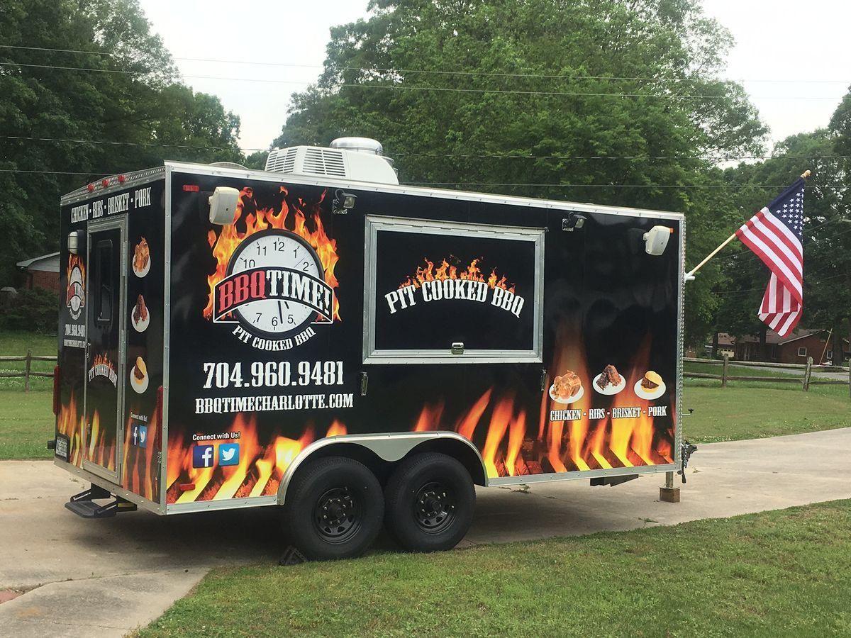 BBQ food truck with flames and American flag parked outdoors beside trees.