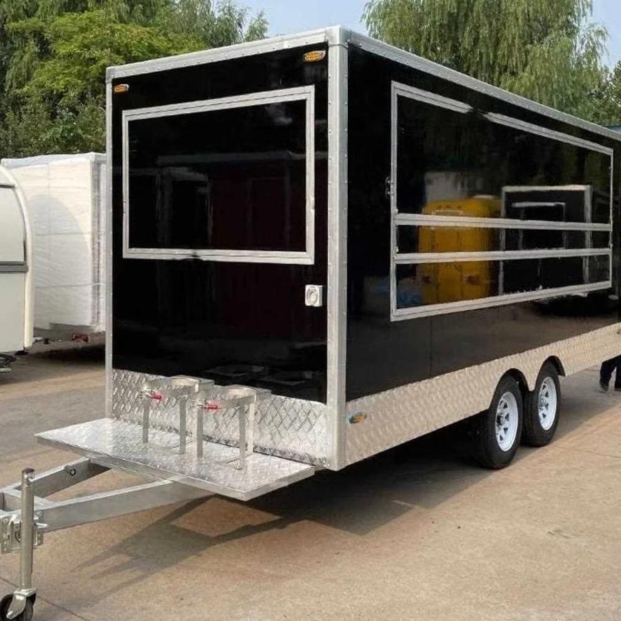 Black trailer with silver trim and dual wheels parked on a concrete surface, surrounded by trees.